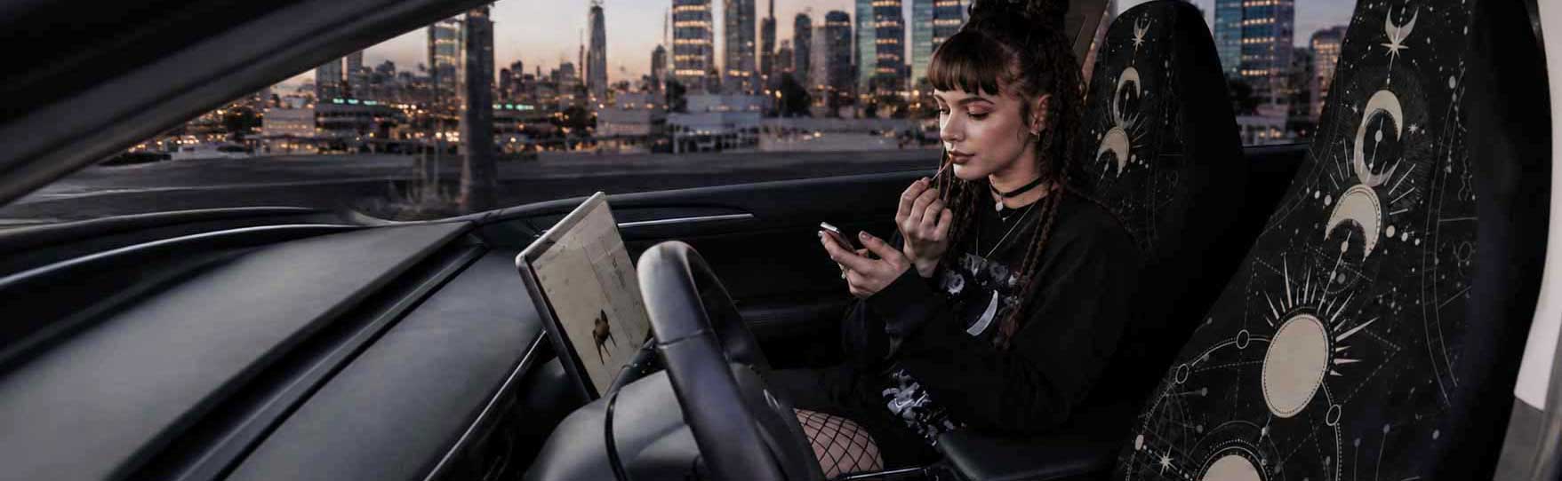 A gothic woman putting makeup on in a car with celestial car seat covers with a cityscape background